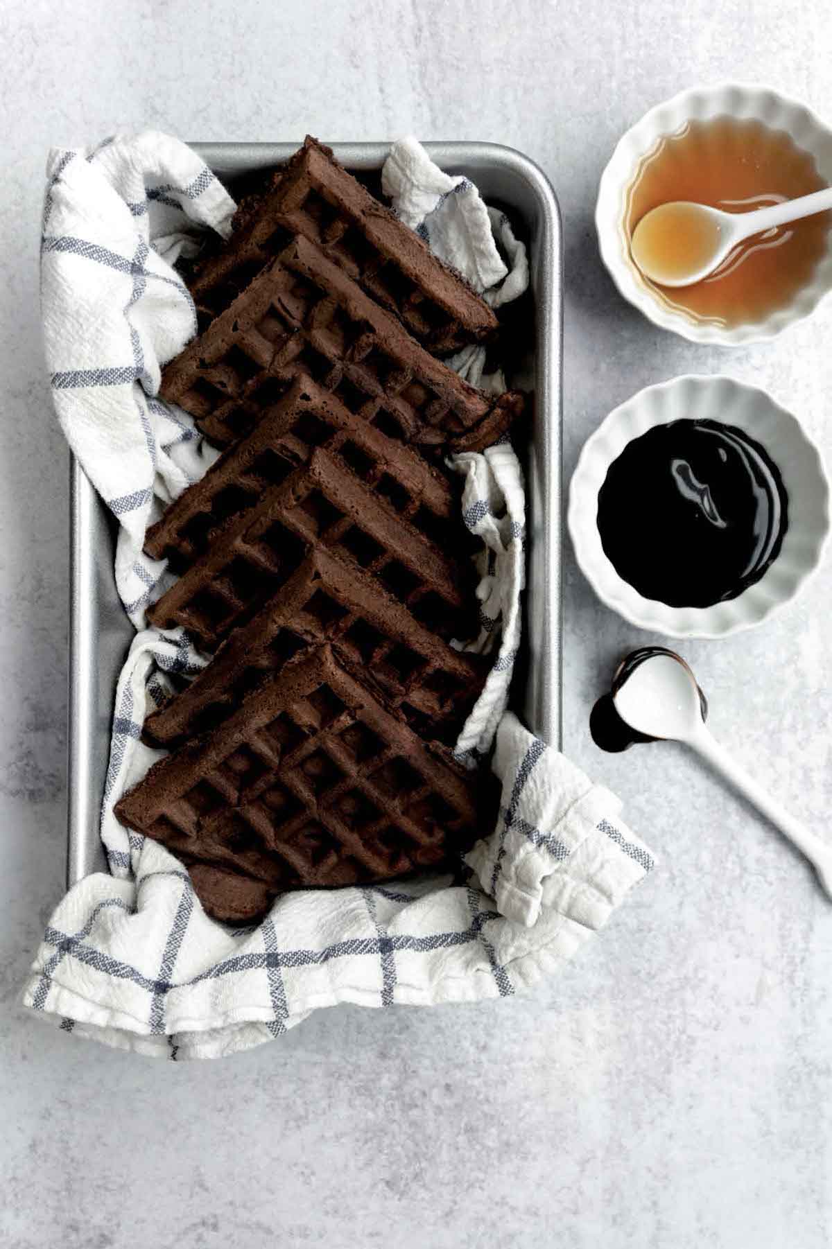 Six square Chocolate Waffles sit in a baking tray next to ramekins of fudge and caramel.