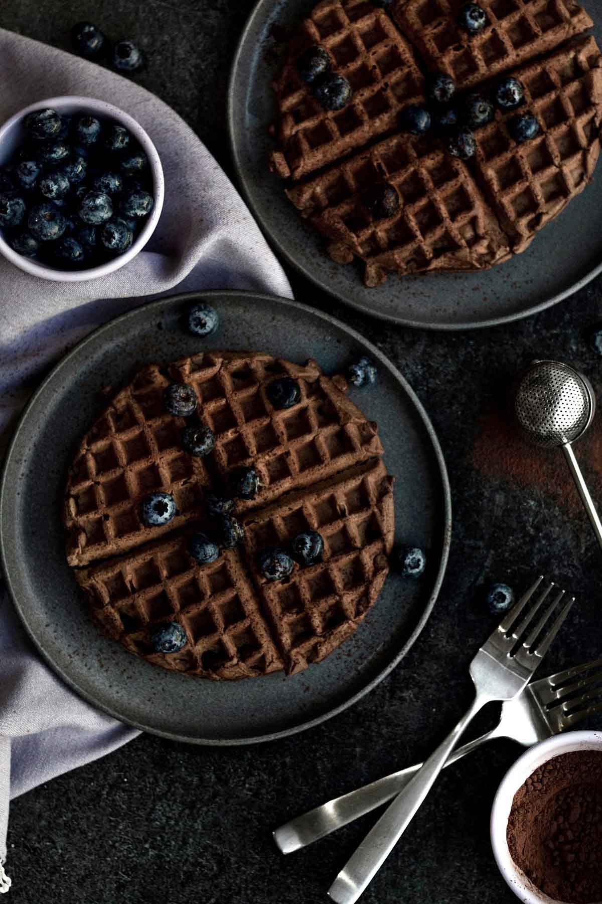 Two circular gluten free Chocolate Waffles on individual black plates topped with blueberries.